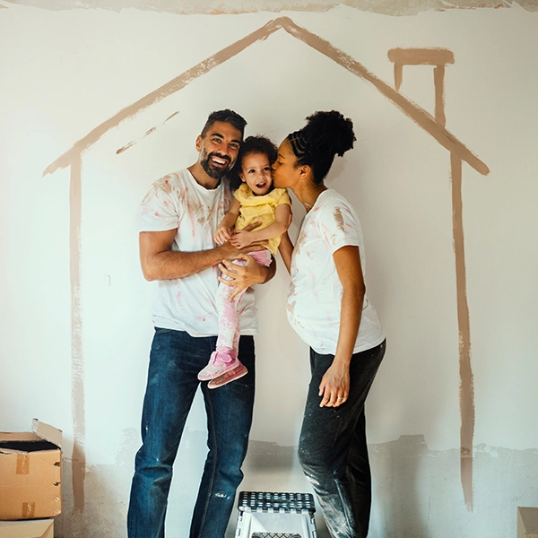 couple hold child while standing in front of a wall with the cutout of a house painted behind them